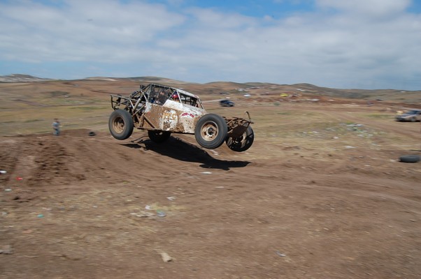 Rusty off-road buggy mid-air, white body with red accents, exposed roll cage, dirt track