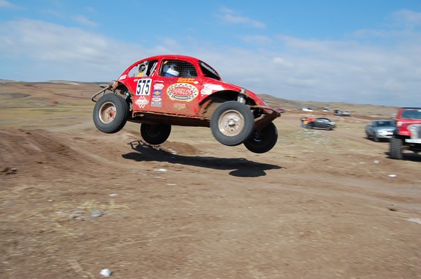 Red off-road Volkswagen Beetle with number 575 airborne over dirt track. Other vehicles visible in background