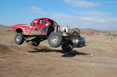 1992 Ford Explorer - A red off-road truck is airborne during a race, with other vehicles and spectators in the background.