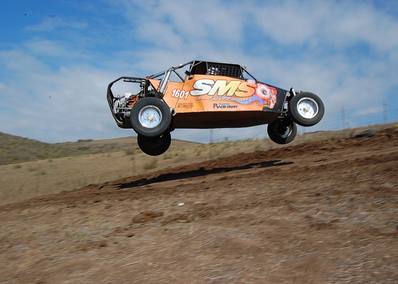 Orange off-road buggy with SMS logo airborne over dirt track