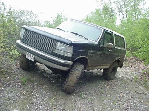 1993 Ford Bronco - A black Ford Bronco navigates a muddy, wooded trail with its front-left tire elevated.