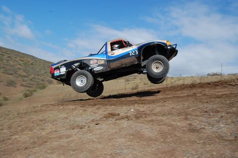 1992 Ford Explorer - A modified truck with racing decals takes flight over a dirt track under a clear blue sky.