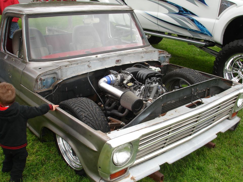 Customized pickup truck with engine exposed, showing aftermarket intake and exhaust. Another lifted truck is visible behind it