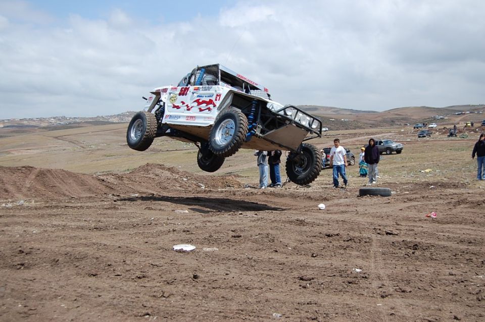 Off-road buggy mid-air jump over dirt track, spectators watch. Other vehicles in background