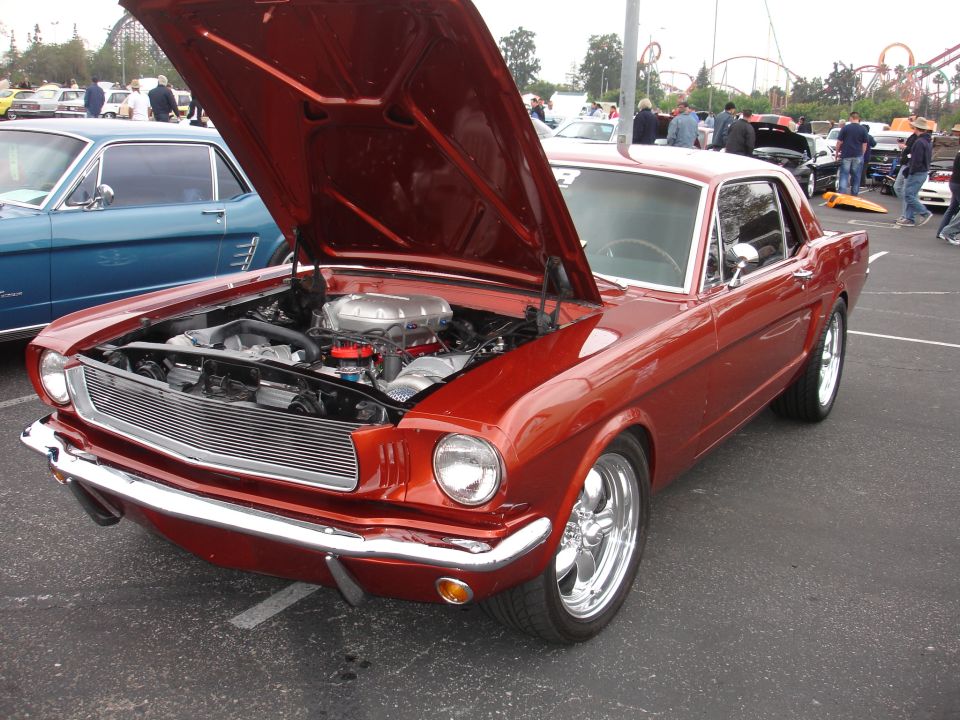 Red 1965 Ford Mustang with hood open, showing engine. Another blue classic car is parked next to it