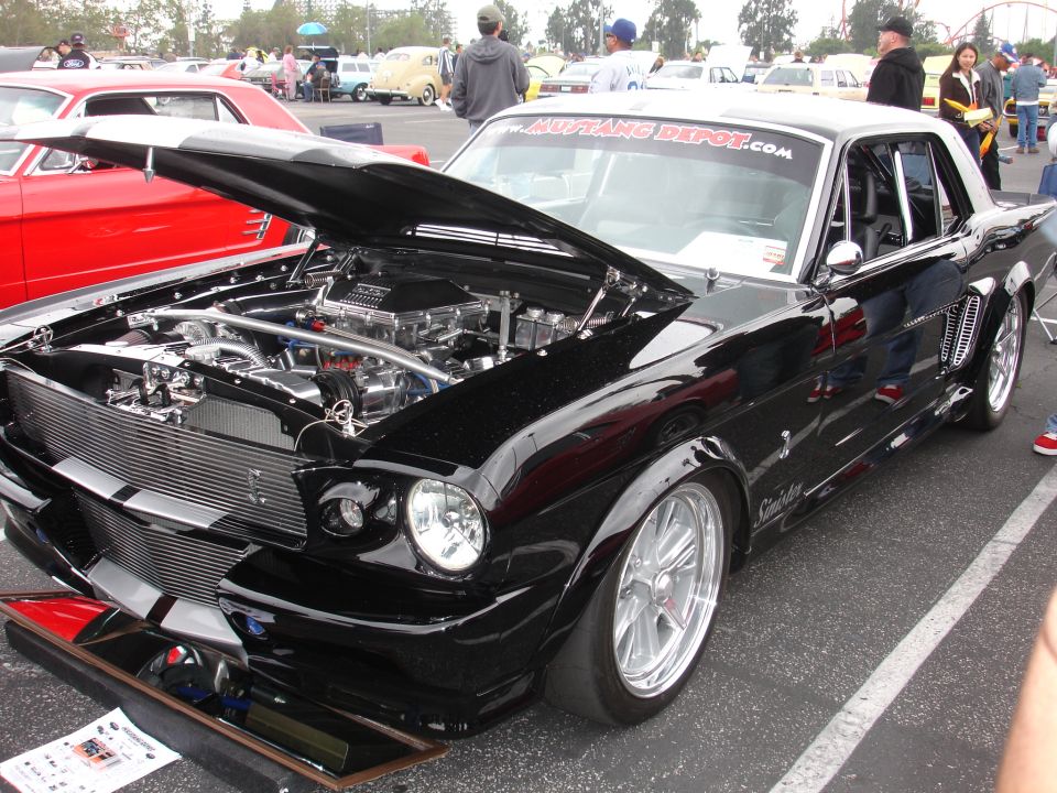 Black modified 1965 Ford Mustang with hood open showing engine, parked next to a red car