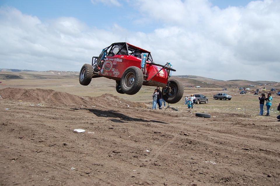 red off-road buggy mid-air over dirt track, people watching in background