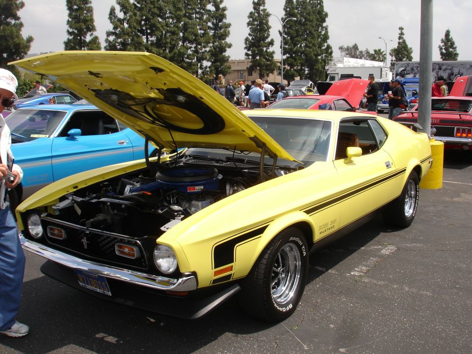 Yellow 1971 Ford Mustang Mach 1 with hood open, showing engine. Another blue car is parked next to it