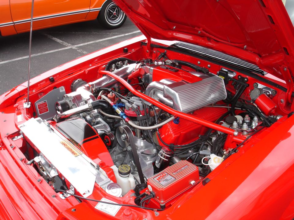 Engine bay of a red car, showing a V8 engine with aftermarket parts like an MSD ignition box and a polished intake. Another orange car is visible in the background