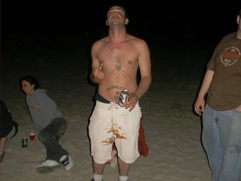 2005 Ford Excursion - A man stands on a beach at night, showing off tattoos and stained shorts while holding a can.