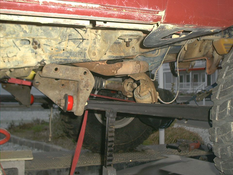 Red vehicle undercarriage with suspension components and driveshaft visible, supported by a jack