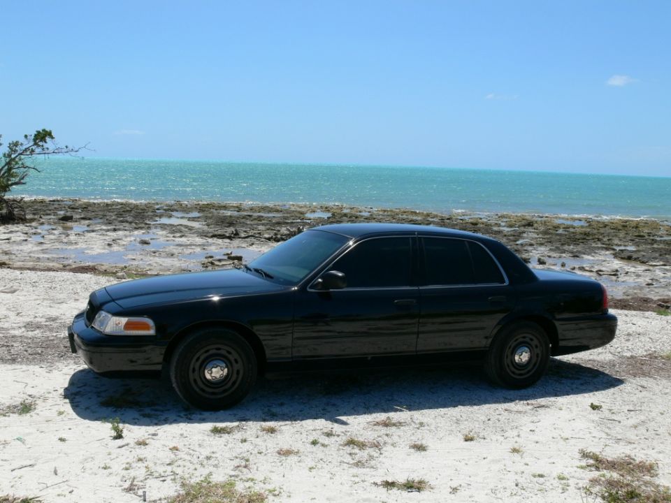 Black Crown Victoria sedan with steel wheels parked on a sandy beach beside turquoise ocean