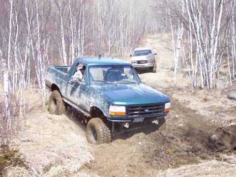 1996 Ford Bronco - Two off-road vehicles navigate a muddy trail through a forest with bare trees.