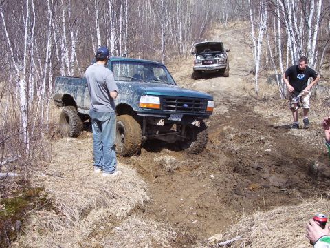 1996 Ford Bronco - Two trucks navigate a muddy off-road trail with trees on either side.