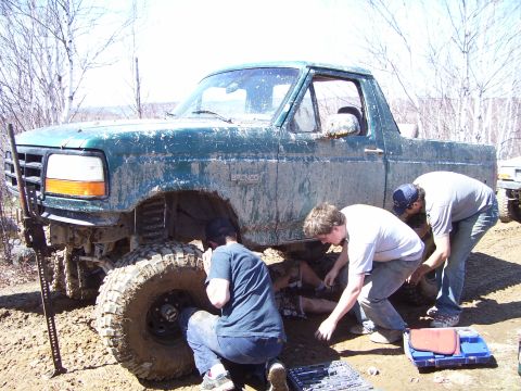 1996 Ford Bronco - Three individuals are gathered around a muddy green Ford Bronco, appearing to perform repairs or modifications on its lifted suspension.