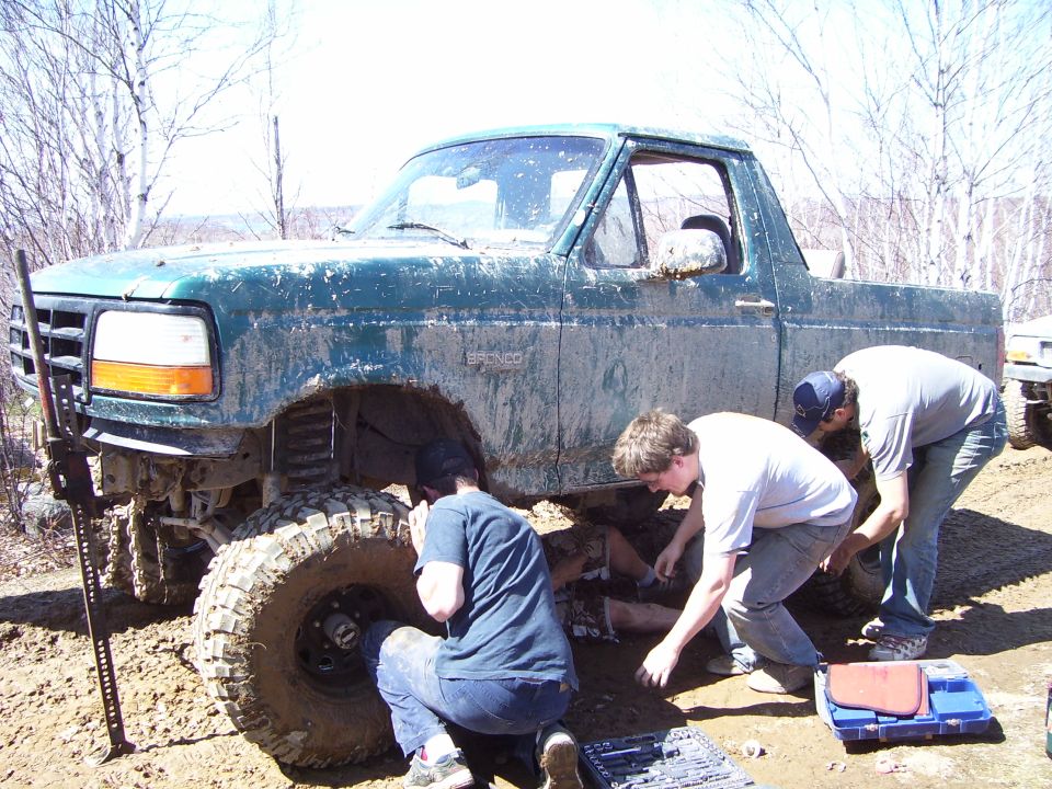 Mud-covered green Ford Bronco with lifted suspension, being worked on by three people outdoors