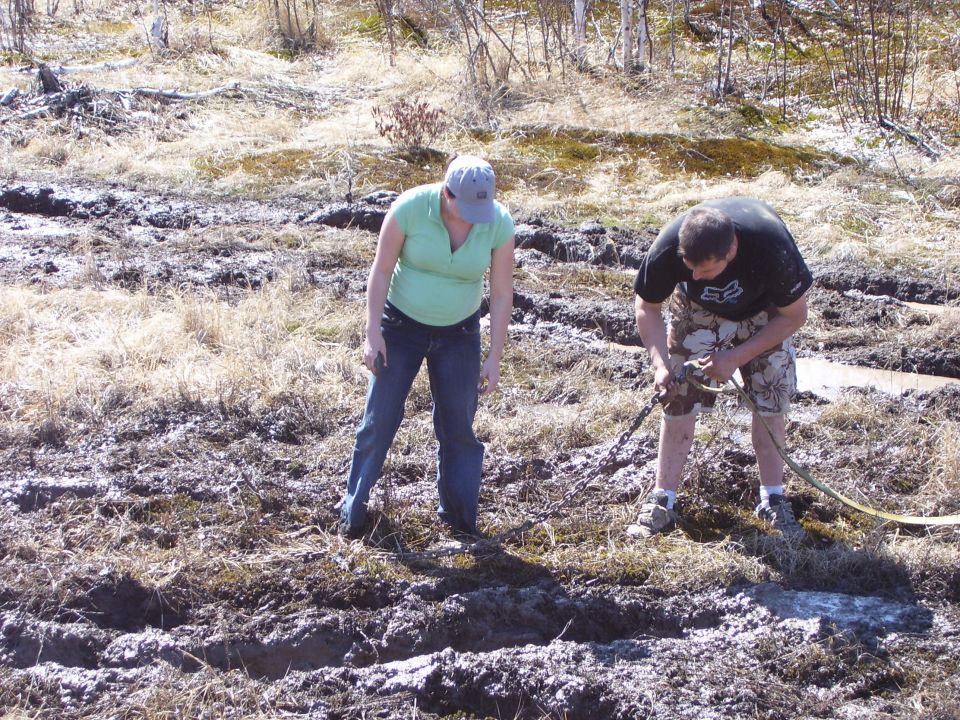 Two people are in a muddy, grassy field. One person is attaching a tow strap to something off-camera