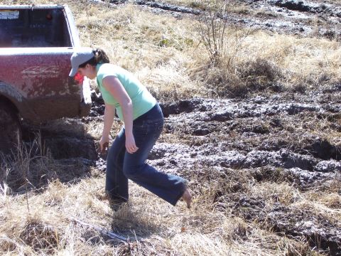 1996 Ford Bronco - A woman stands near a red pickup truck that is stuck in a muddy, grassy field.