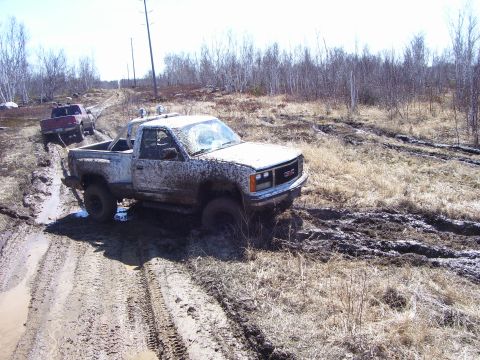 1996 Ford Bronco - Two trucks navigate a challenging muddy off-road course on a bright, overcast day.