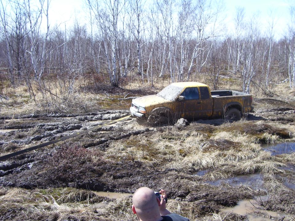 Yellow pickup truck stuck in mud, covered in dirt, with trees in the background