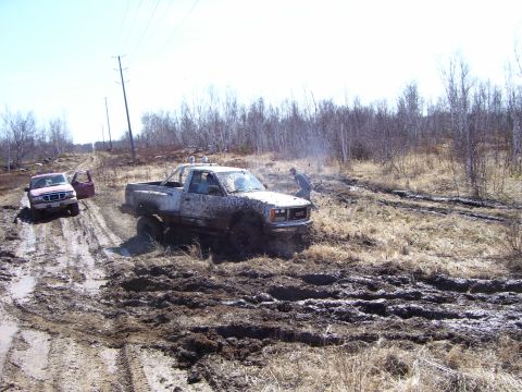 1996 Ford Bronco - Two trucks navigate a challenging muddy terrain, with one GMC pickup appearing stuck in the mire.