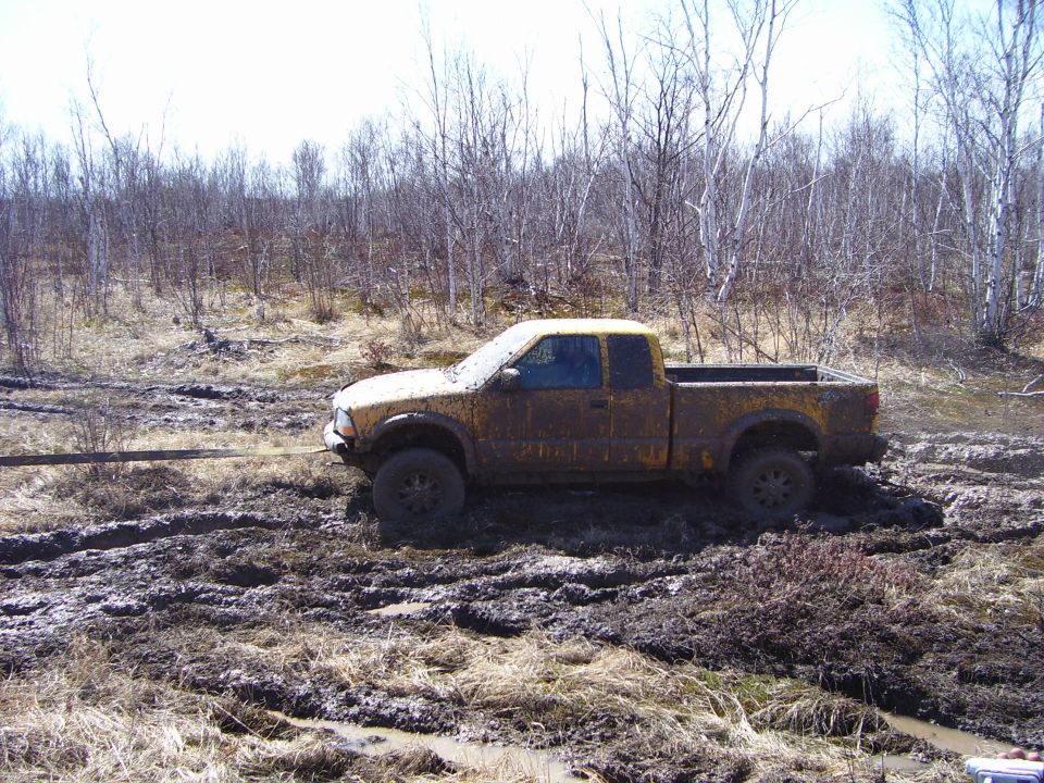 Muddy yellow pickup truck stuck in a muddy field with trees in the background