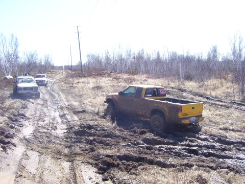 1996 Ford Bronco - Several vehicles navigate a challenging muddy off-road course under a bright sky.