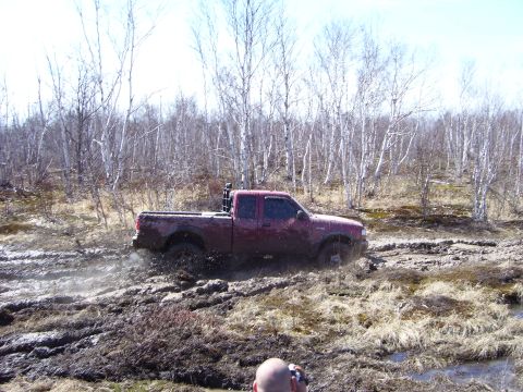 1996 Ford Bronco - A red pickup truck navigates a muddy off-road course, kicking up mud as it struggles through the terrain.
