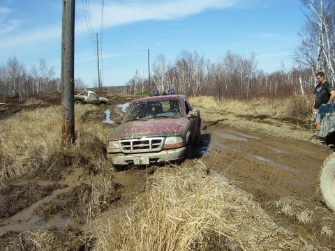 1996 Ford Bronco - Two trucks are stuck in a muddy off-road area under a clear blue sky.