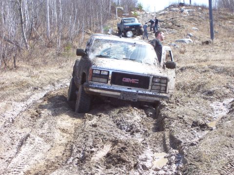 1996 Ford Bronco - A GMC truck is stuck deep in the mud on an off-road trail, with another vehicle and people visible further up the hill.
