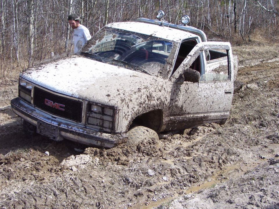 Muddy GMC pickup truck stuck in a muddy ditch with a person standing nearby