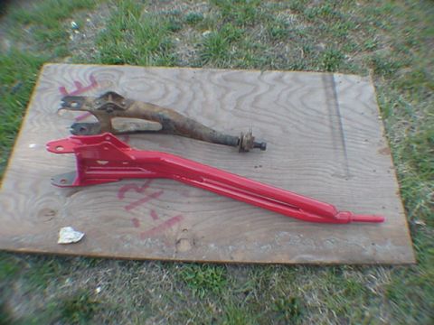 1993 Ford Bronco - Two suspension control arms, one red and one rusty brown, are displayed on a plywood board outdoors.