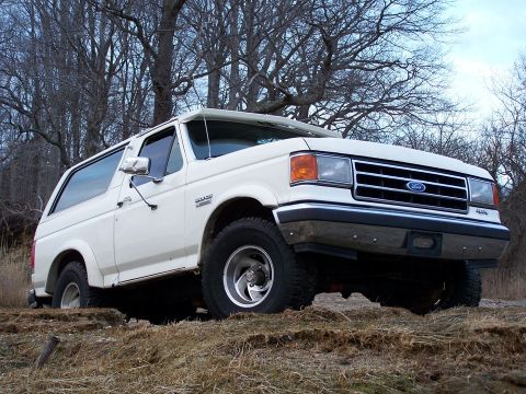 1989 Ford Bronco - A white Ford Bronco is parked on a dirt hill with trees in the background.