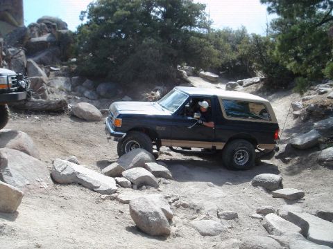 1994 Ford Bronco - A Ford Bronco drives over rocks on an off-road trail, with another vehicle partially visible to the left.