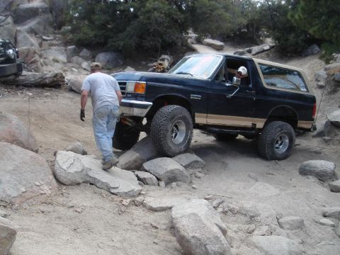 1994 Ford Bronco - Dark blue two-door Ford Bronco with tan lower two-tone and tan hardtop straddling boulders on a rocky trail while a man in a gray shirt and…