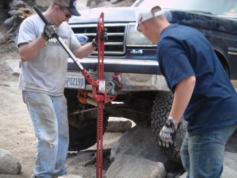 1994 Ford Bronco - Two men in gloves work a red Hi-Lift jack at the front bumper of a dark blue Ford 4x4 on a rocky trail, one steadying the jack handle while…