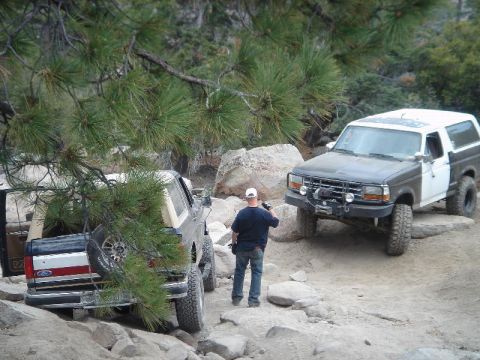 1994 Ford Bronco - Two full-size Ford Broncos on a rocky pine-lined trail, the near one dark with a tan top and a spare wheel carried on the rear, the far one…