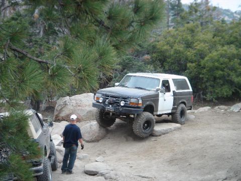 1994 Ford Bronco - Dark green and white two-door Ford Bronco flexing over a rock ledge on a pine-lined dirt trail, front tire lifted onto granite. A winch-equ…