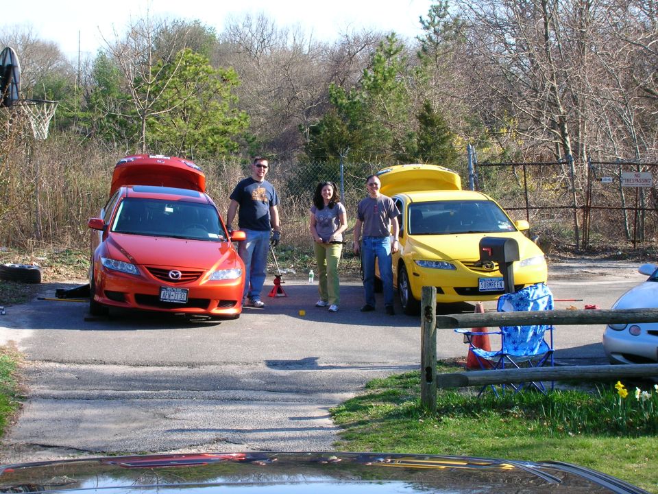 Two people stand between an orange and a yellow Mazda 6, both with trunks open