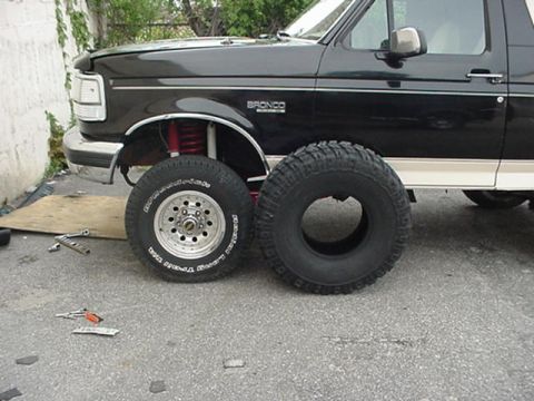 1993 Ford Bronco - Driver-side front of a black Ford Bronco with tan lower two-tone and BRONCO fender badging, jacked up in a driveway with red coil springs a…