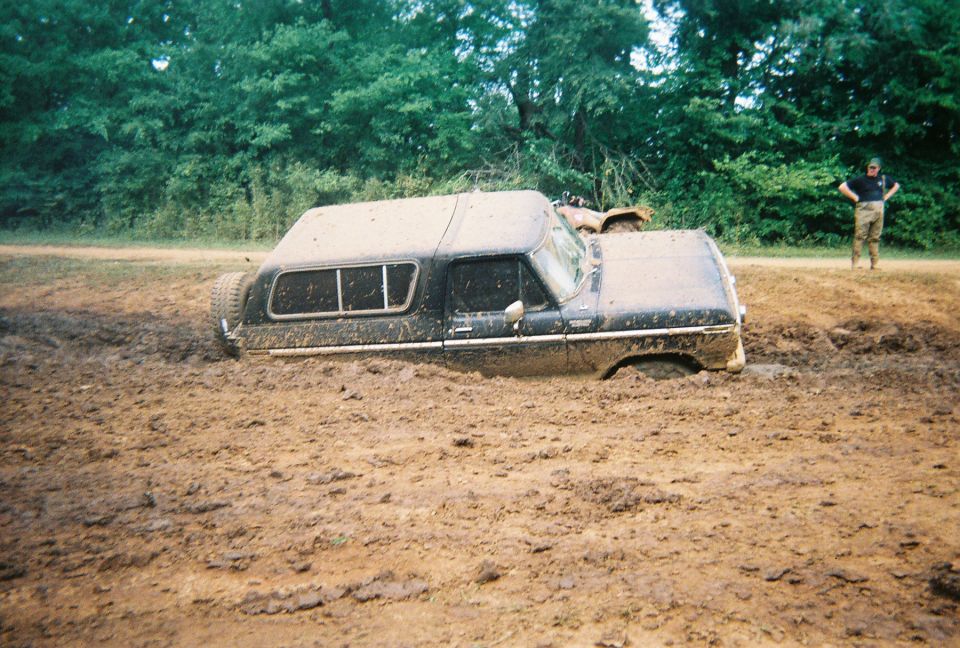 Mud-covered black 1978 Ford Bronco with a camper shell stuck in deep mud, viewed from the front-right