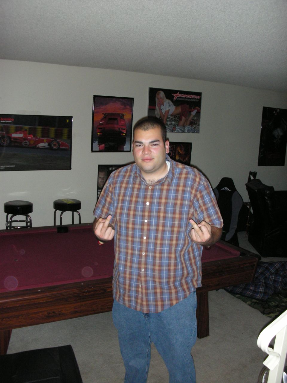 Guy in plaid shirt and jeans stands in front of a pool table, giving the finger. Posters of cars and women are on the wall behind him