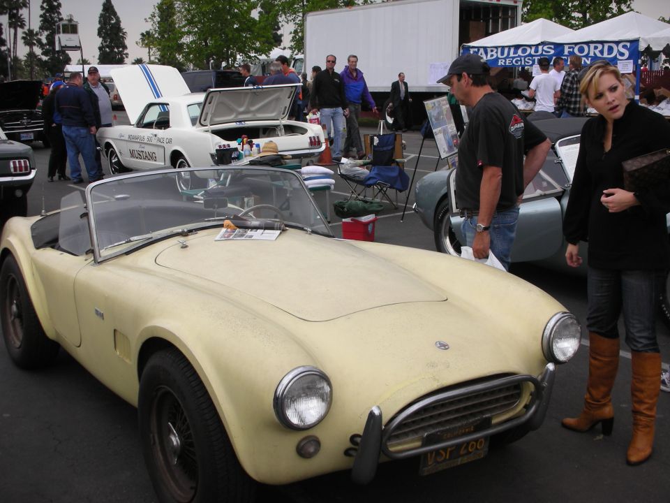 Yellow AC Cobra replica with wire wheels and chrome bumper. A white Mustang is visible behind it