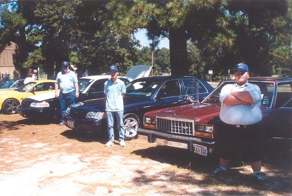 Row of sedans parked under trees with three men standing nearby at an outdoor gathering