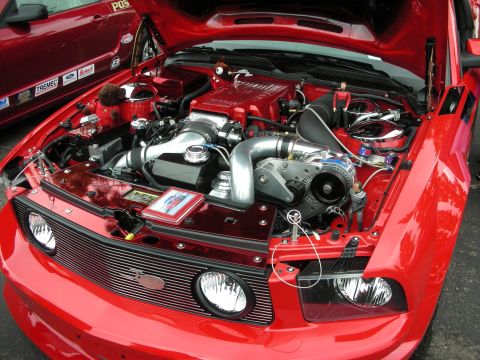 1992 Ford Crown Victoria - The engine bay of a red Ford Mustang, modified with a supercharger, is displayed with two small figurines placed among the components.