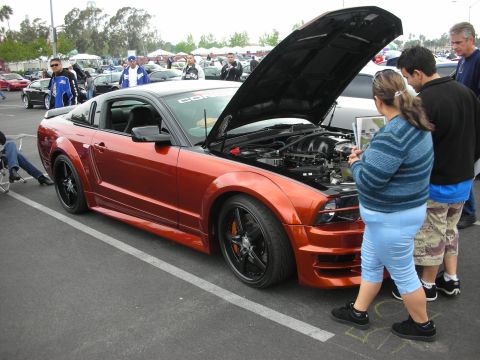 1992 Ford Crown Victoria - Orange Ford Mustang at a car show with its hood open, drawing attention from onlookers.