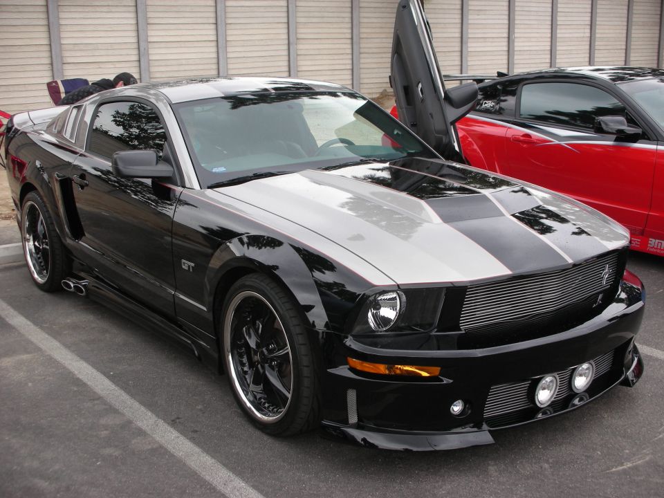 Black Ford Mustang GT with silver racing stripes and lambo doors, parked next to a red car