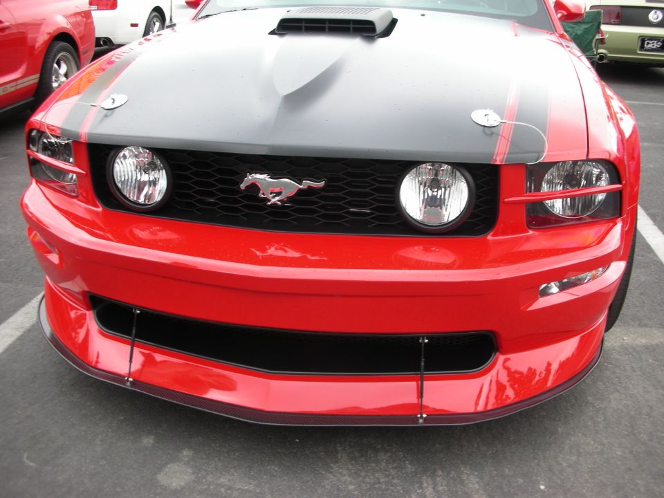 Red Ford Mustang front with black hood stripes and a running horse emblem on the grille. Another red car is visible to the left