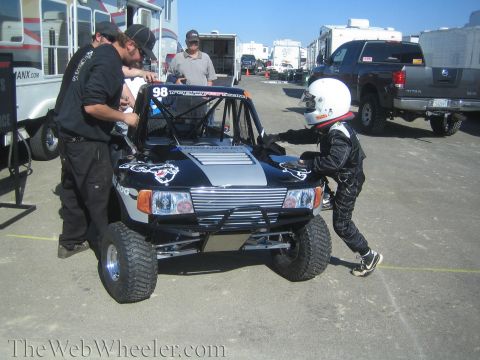 1990 Ford Bronco - driver in a white helmet and black suit climbs into a black No. 98 off-road race truck while crew work nearby in a paddock.