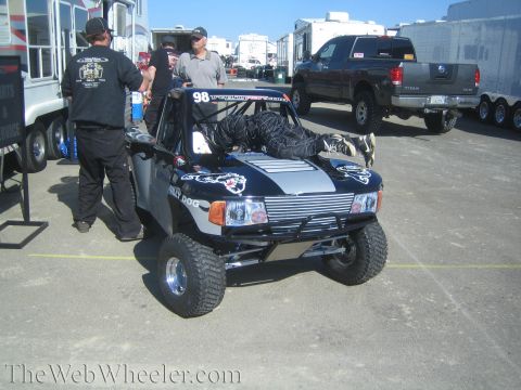 1990 Ford Bronco - black No. 98 single-seat off-road race truck sits in a paddock with its driver reclined inside and crew standing alongside.